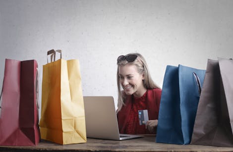 Woman seated browsing her laptop with shopping bags on the table, looking into ecommerce loans for her online business